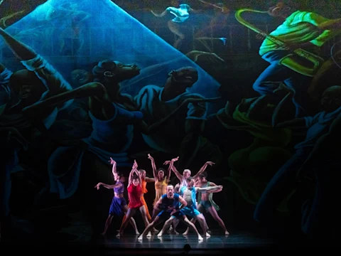 A group of dancers in colorful outfits perform on stage in front of a large, dramatic mural depicting expressive figures in blue and green tones.
