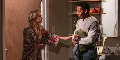 A woman in a robe gestures toward a man holding clothing in a kitchen area, both appearing engaged in a serious conversation.