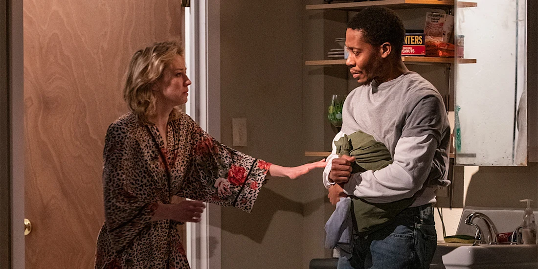 A woman in a robe gestures toward a man holding clothing in a kitchen area, both appearing engaged in a serious conversation.
