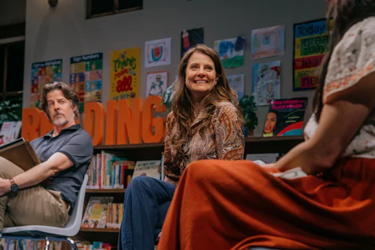 Three adults sit and talk in a library, surrounded by books and colorful children's artwork; large orange letters spell "READING" in the background.