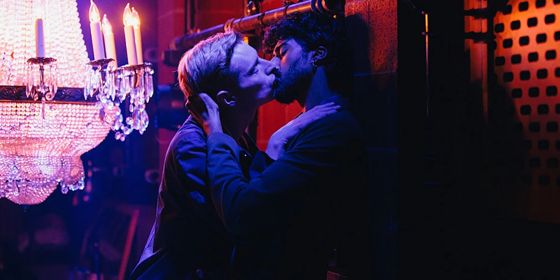 Two men are kissing passionately in a dimly lit room with colorful lighting and a chandelier.