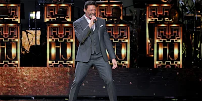 A man in a gray suit sings into a microphone on stage with an art deco inspired backdrop and illuminated band stands behind him.