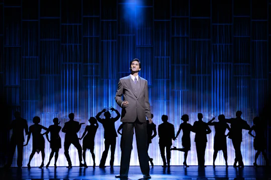 A man in a gray suit stands center stage under a spotlight, with a blue-lit backdrop and silhouettes of people posing behind him.