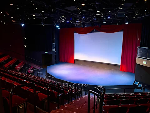 Empty theater with red seats and a large stage framed by red curtains and a blank white screen, viewed from the audience.