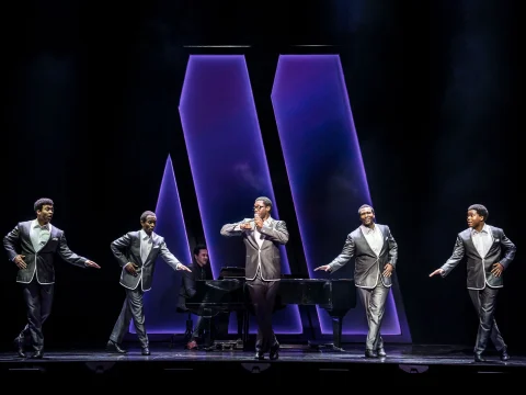 Five performers in matching suits are singing and dancing on stage in front of a pianist, with large purple-lit shapes in the background.