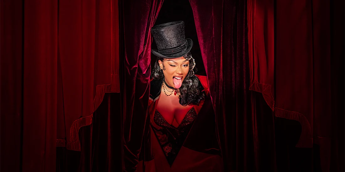 A person wearing a black top hat and dark corset-style outfit opens red velvet curtains, sticking out their tongue and smiling.