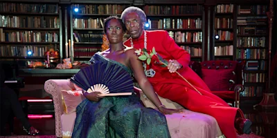 Two people pose on an ornate couch in a library; one in a green dress holding a fan, the other in a bright red suit holding a rose. Books line the shelves in the background.
