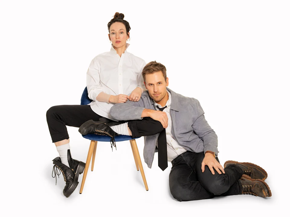 A woman sits on a chair wearing a white shirt and black pants, while a man in a gray jacket and black pants sits on the floor, leaning against her.