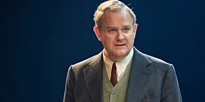 A middle-aged man in a suit, tie, and vest stands against a dark blue background, looking slightly to the side with a neutral expression.