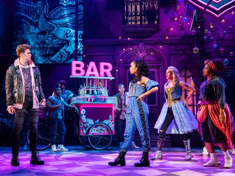 A group of actors on stage perform in a colorful, vibrant set with a neon "BAR" sign, dressed in modern and historical costumes, featuring a bar cart in the background.