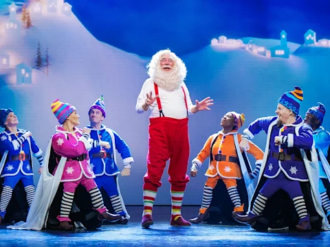 A man dressed as Santa Claus stands center stage, surrounded by performers in colorful elf costumes against a snowy, winter village backdrop.
