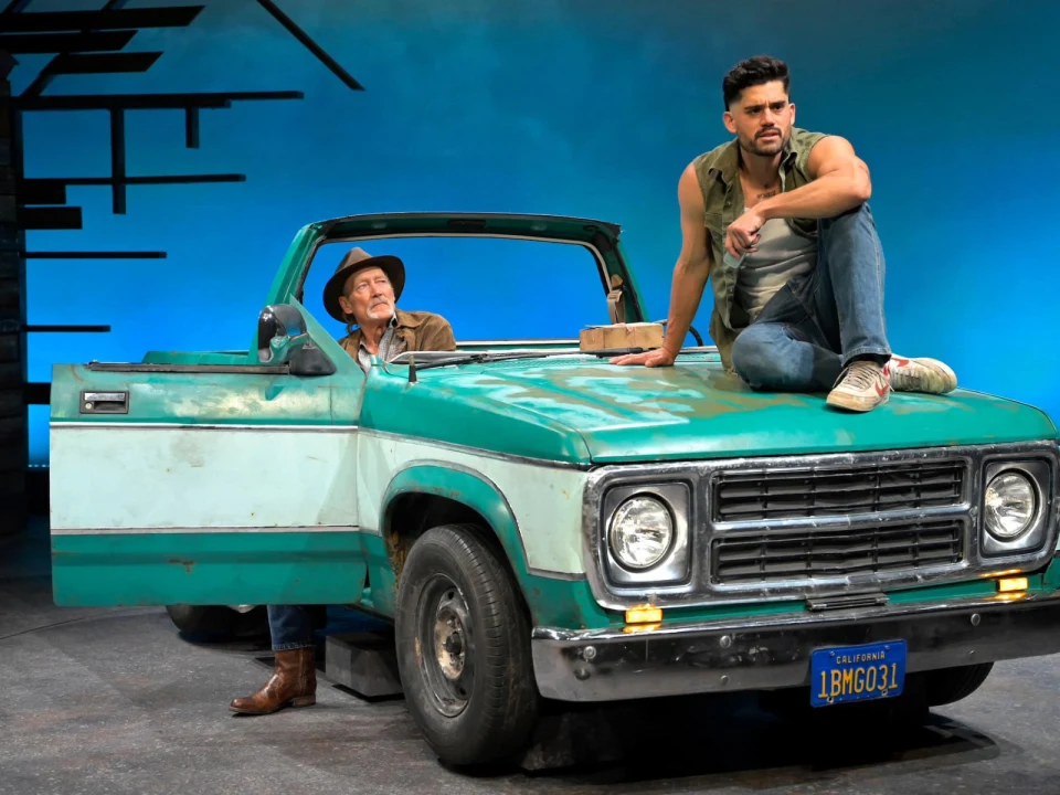  Production photo of Mother Road in California, with Emilio Garcia-Sanchez as Martín Jodes and James Carpenter as William Joad on an old, weathered pickup truck on a stage.