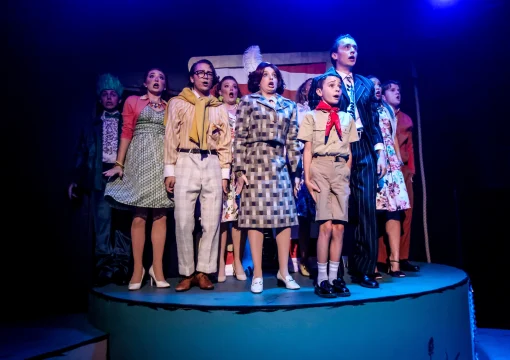 A group of actors on stage, standing close together in costume under bright stage lighting, appear to be singing or speaking in unison. The background is dark with hints of a set design.