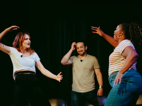 Three people are standing on stage performing. One woman raises her arms, a man scratches his head, and another woman gestures with her hands raised. All are wearing microphones.