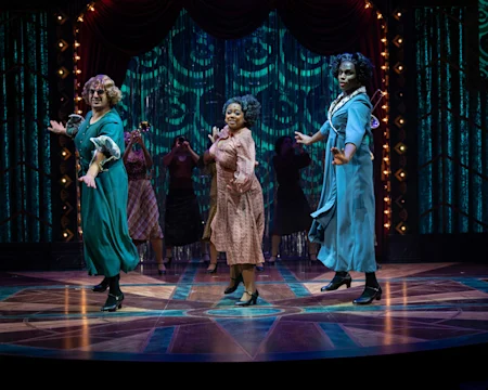 Three performers in vintage costumes dance on a theater stage with dramatic lighting and a background of sparkling blue curtains and red drapes.