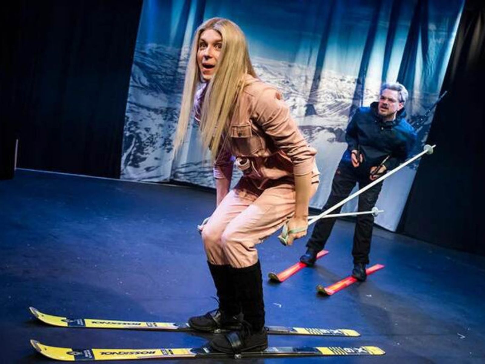 Two people on stage in skiing poses, one in front wearing a pink outfit and the other behind in dark clothing, with a snowy mountain backdrop.