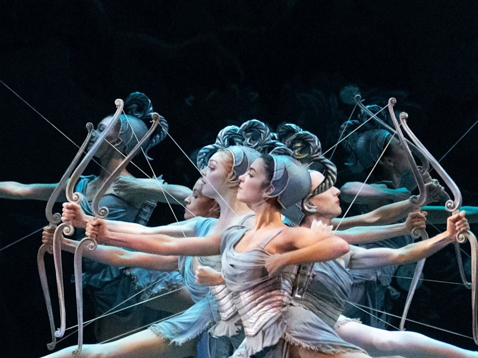 Four ballet dancers dressed in gray costumes pose with bows, standing in a line with arms extended, creating a symmetrical and dynamic visual effect on stage.