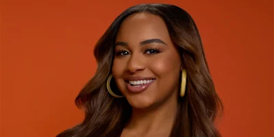 A woman with long brown hair, wearing large hoop earrings, smiles against an orange background.
