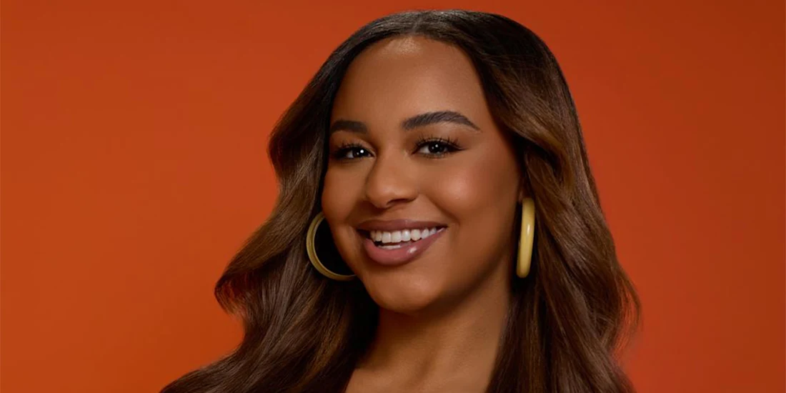 A woman with long brown hair, wearing large hoop earrings, smiles against an orange background.