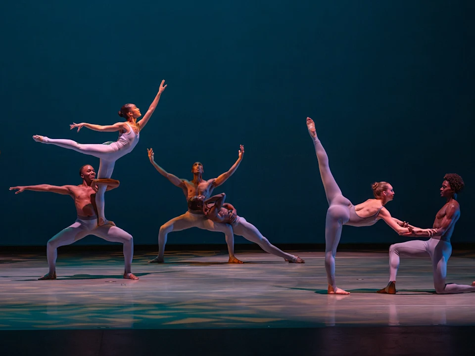 Six dancers in white costumes perform on stage, with three pairs in dynamic poses; one woman is lifted, another arches back, and one extends a leg high. Blue-lit backdrop.