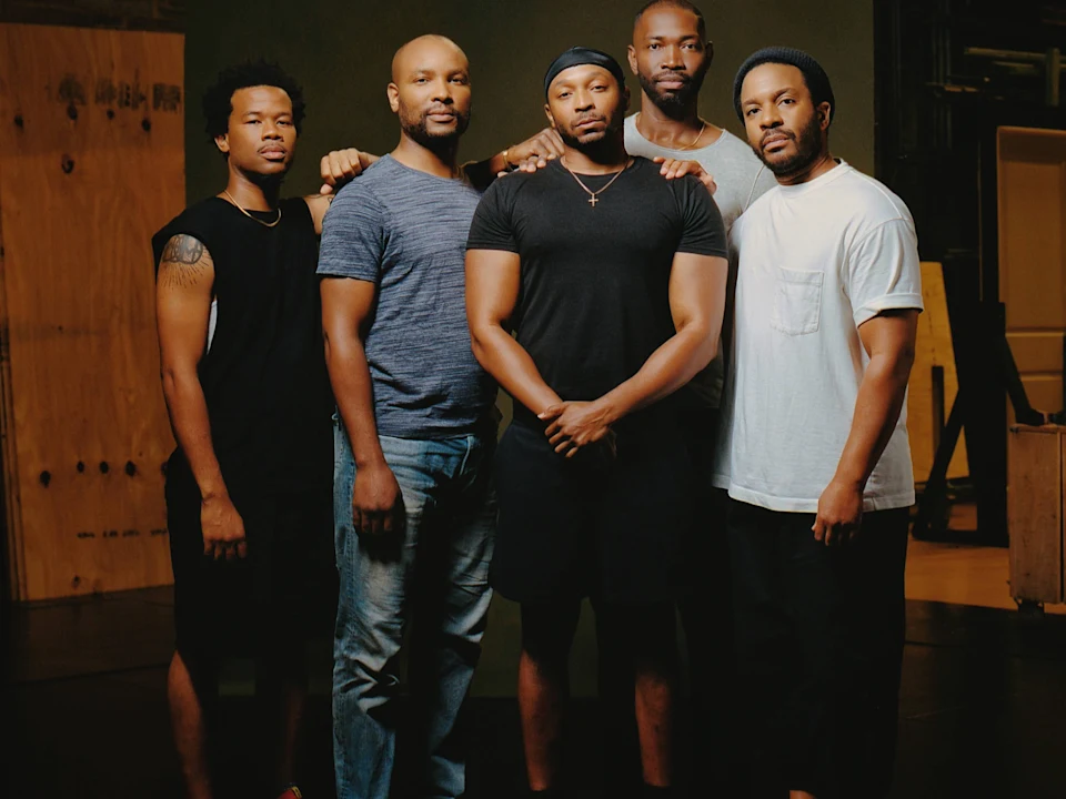 Five men stand side by side indoors, facing the camera, with neutral expressions and casual clothing.