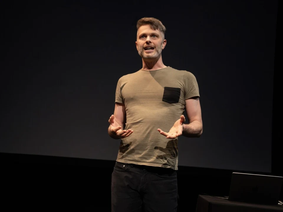 A man in a t-shirt speaks on stage in front of a dark background, with a laptop on a nearby table.