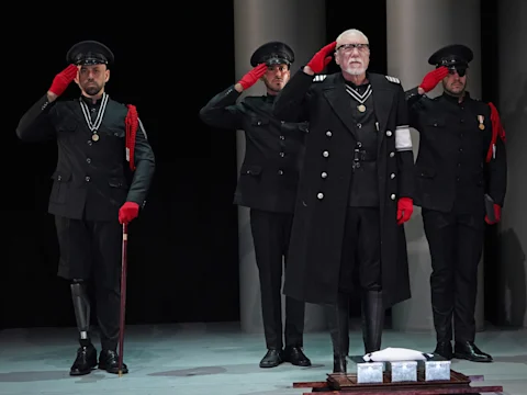 Four men in black military-style uniforms and red gloves stand on stage, saluting. The man in front stands on a platform; two have prosthetic legs. The backdrop is plain with columns.