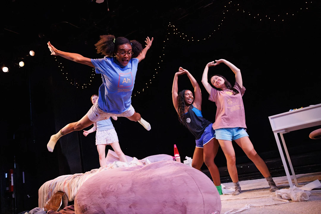 Four young people in casual clothes are on stage; one is jumping onto a bed while the others stand nearby, smiling and posing with arms up. String lights hang in the background.