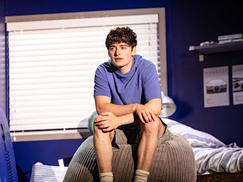 A teenage boy perches on a beanbag in his bedroom, looking up mid-conversation.