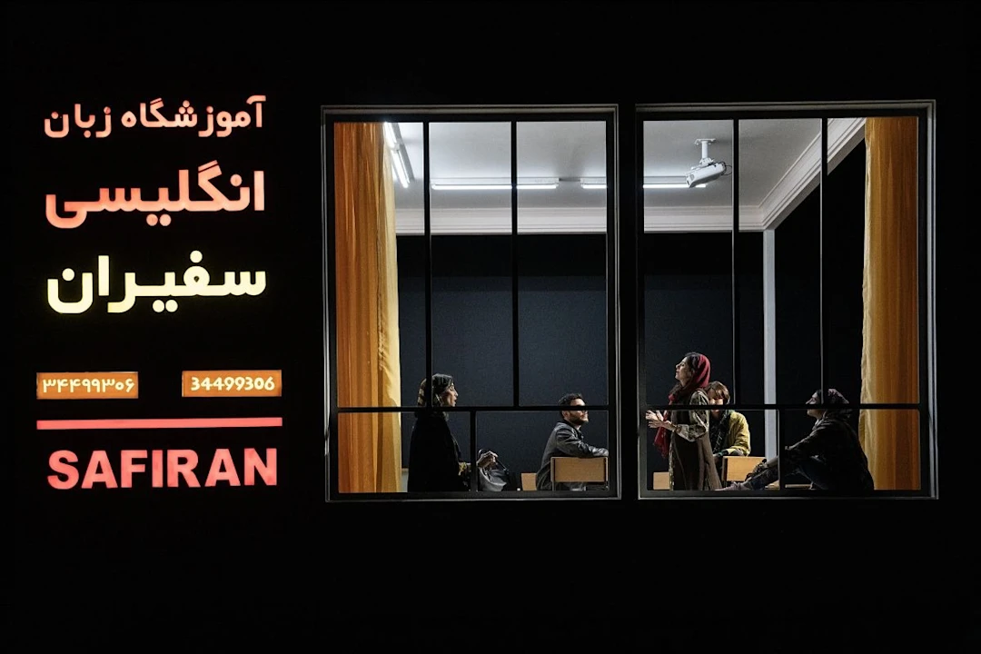 A group of people sit and converse in a classroom visible through large windows at night; illuminated signage outside reads "SAFIRAN" in English and Farsi.