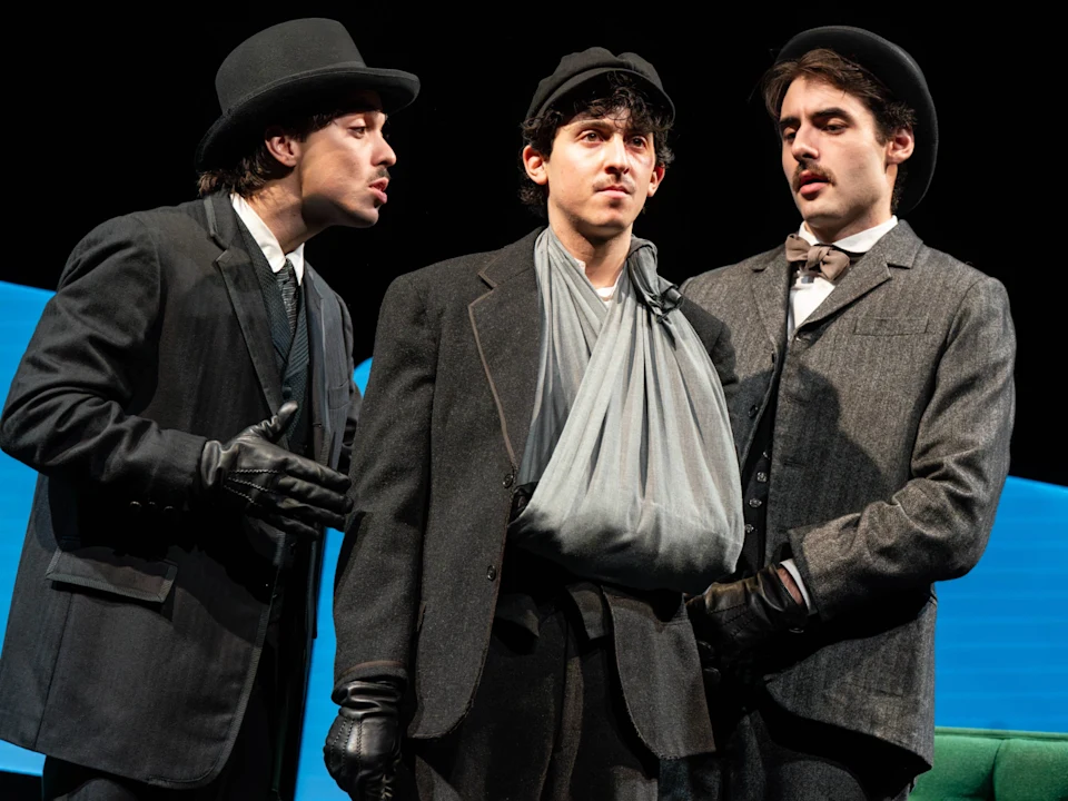 Three men in early 20th-century attire stand together; the center man has his arm in a sling, while the other two appear to be speaking to him.
