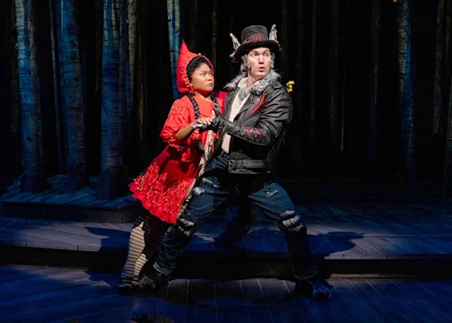 Two actors on stage: one dressed as Little Red Riding Hood in red, and the other as the Wolf in a leather jacket and fur, performing in a forest set.