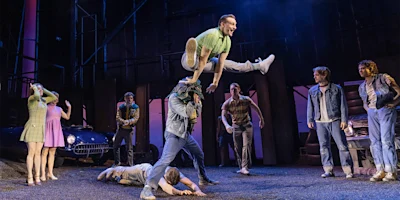 A man in a green shirt leaps over another man's shoulders onstage, surrounded by other actors in 1950s-style costumes, with a vintage car in the background.