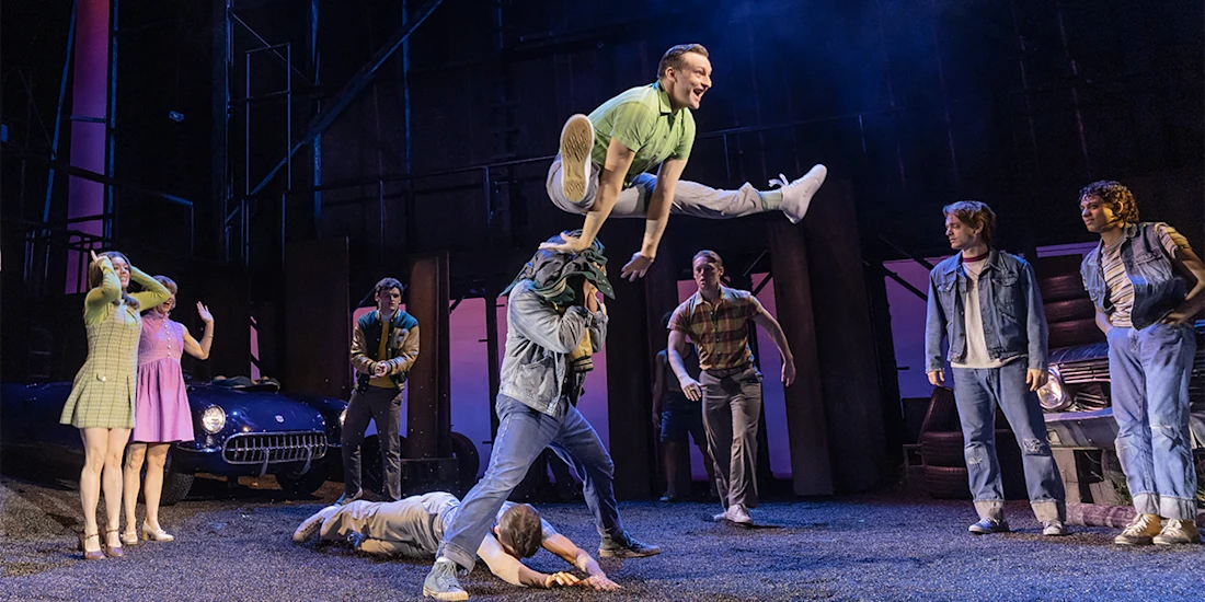 A man in a green shirt leaps over another man's shoulders onstage, surrounded by other actors in 1950s-style costumes, with a vintage car in the background.