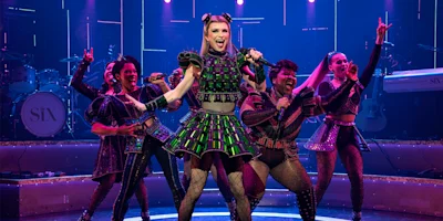 Five performers in colorful, Tudor-inspired costumes sing and dance energetically on a stage with band instruments and pink-purple lighting.