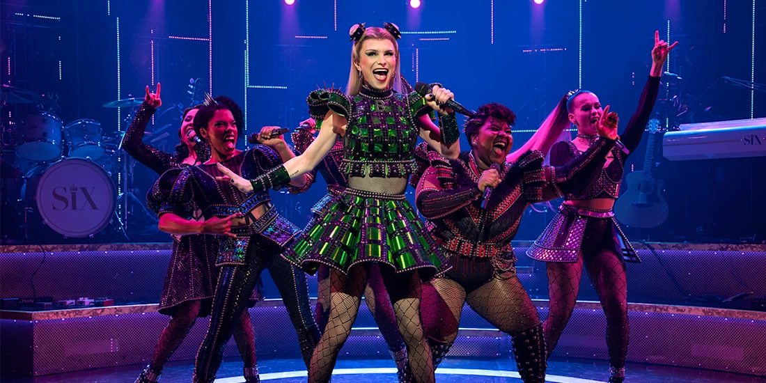 Five performers in colorful, Tudor-inspired costumes sing and dance energetically on a stage with band instruments and pink-purple lighting.