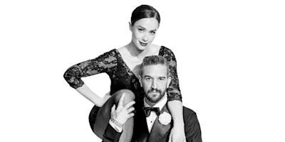 A woman in a black lace dress sits with one arm resting on the shoulder of a seated man in a tuxedo against a plain white background.