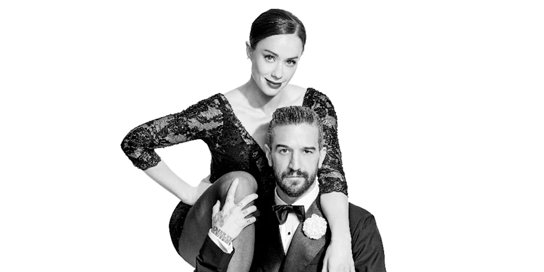 A woman in a black lace dress sits with one arm resting on the shoulder of a seated man in a tuxedo against a plain white background.