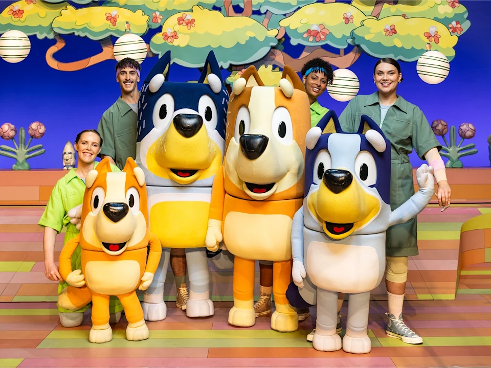 Four people in green costumes stand on a colorful stage with four large mascot costumes of Bluey characters in front of a cartoon-like backdrop with trees and lanterns.