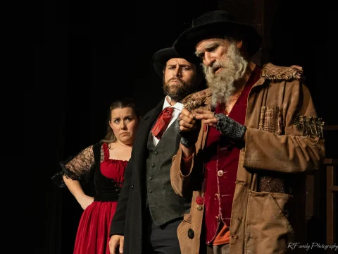 Three actors in period costumes performing on stage, with a woman in a red dress, a man in a suit and hat, and an older man with a gray beard and tattered coat.