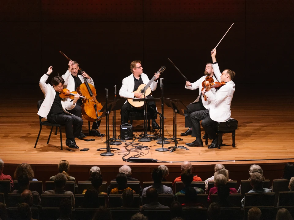 A musical quintet dressed in white jackets performs on stage with a guitar and string instruments. An audience watches the performance from their seats.