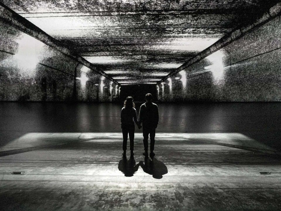 A couple holds hands while standing in a dimly lit tunnel, casting shadows on the ground.