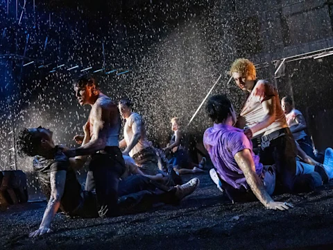A group of men, some shirtless and others in wet clothing, engage in a dramatic, rain-soaked fight scene on a dark stage.
