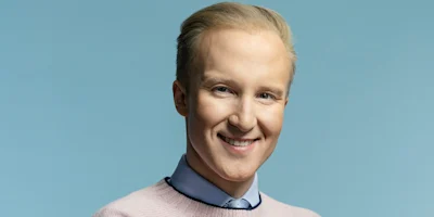 A person with short blond hair, wearing a light blue collared shirt, tie, and pale pink sweater, smiling against a plain blue background.