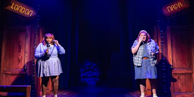 Two performers stand on a stage under purple lighting, each holding a microphone. Signs above read "London" and "Napa," with wooden panels and a house set piece in the background.