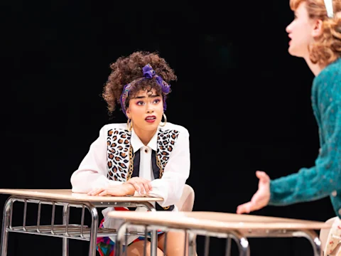 A young woman with curly hair and a headband sits at a desk, looking intently at another person whose arm is extended, suggesting a conversation or discussion.
