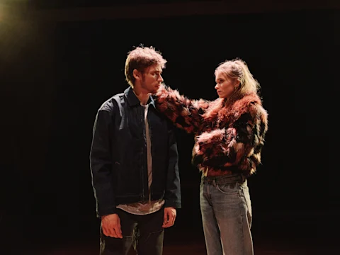A woman in a fur jacket touches a man's chin while facing him. The man wears a denim jacket and looks at her. They stand under a spotlight against a dark background.