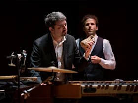 Two men in formal attire perform on stage; one plays percussion instruments and holds a shaker, while the other stands in the background.