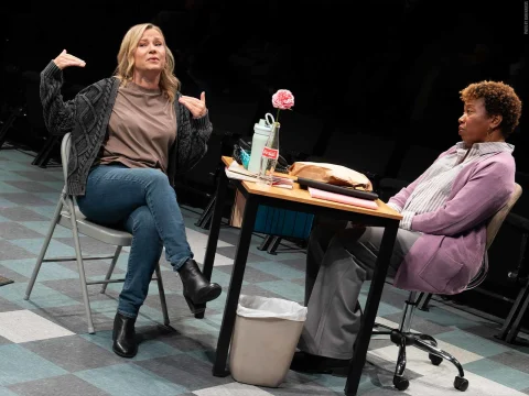 Two women are engaged in a conversation. One is seated on a chair gesturing with her hands, while the other sits at a table with a notepad and a vase with a single flower.