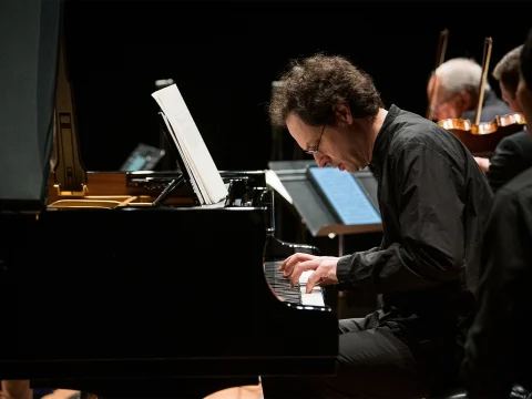 A man in a black shirt plays a grand piano, concentrating on the sheet music in front of him. In the background, other musicians are seated with their instruments.